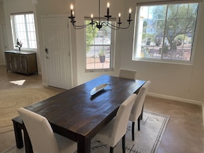 A dining room with a dark wooden table, white upholstered chairs, a chandelier, and large windows with a view of a garden.