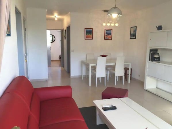 A modern living room with a red sofa, a white coffee table, and a dining area with white chairs and a table. There is a ceiling light fixture above the dining area.