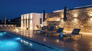 A swimming pool with lounge chairs and umbrellas, a stone wall, and a modern building with lights. - Bracket House Paros Villa with Private Pool Sleeps 8 (Paros)