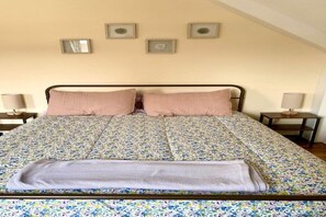 Una cama bien hecha con un edredón floral, dos almohadas y una manta doblada sobre un somier de metal en una habitación con techo inclinado y cuadros en la pared.
