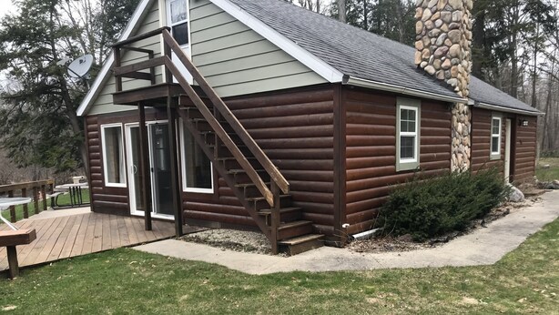A log cabin with a wooden deck, a stone chimney, and a large window. - Riverfront Property Great for Fishing or Just Relaxing (Hesperia)