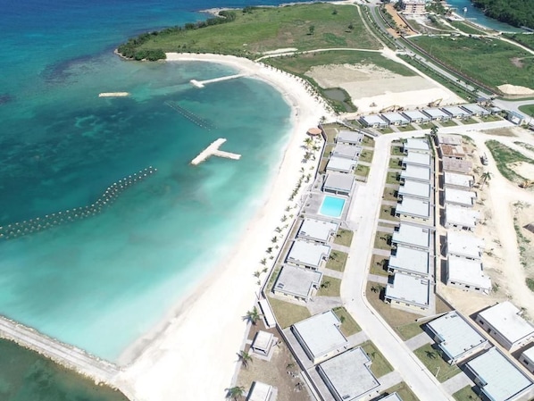 Vue aérienne d’une station balnéaire avec une plage, une jetée et un ensemble de bâtiments modernes dotés d’une piscine.