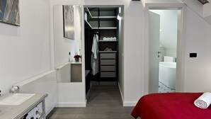 Une chambre à coucher avec une couvre-lit rouge, un radiateur blanc, un miroir et un couloir menant à une salle de bain équipée d’une baignoire.