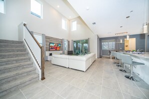A spacious living area with a staircase, a large TV, a glass partition, and a kitchen area with bar stools.