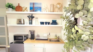 A compact kitchen with white cabinets, a microwave, a coffee maker, and a sink. There are shelves above the counter holding various kitchen items and a potted plant. - Newly Remodeled Studio in between SF and Napa! (Benicia)