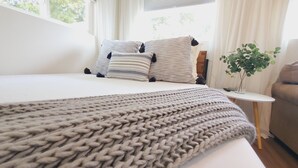 A neatly made bed with a grey knitted blanket, striped pillows, and a small bedside table with a plant. - Newly Remodeled Studio in between SF and Napa! (Benicia)