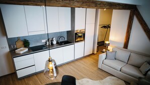 Een moderne keuken met witte kastjes, een zwart aanrecht en een houten plafond. Er is een eethoek met een tafel, stoelen en een schaal fruit. Een bank en een lamp staan in de woonkamer.