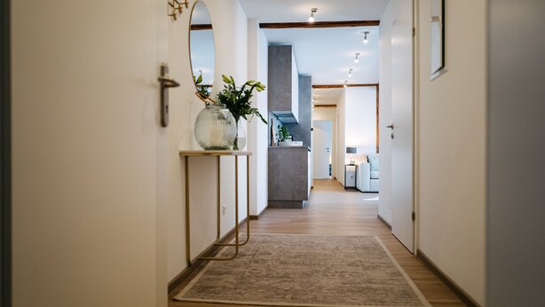 Een gang met een houten vloer, een vloerkleed, een vaas met planten, een spiegel en een deur naar een keuken.
