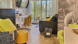 A modern living room with a fireplace, a television, and a large window with a view of trees.