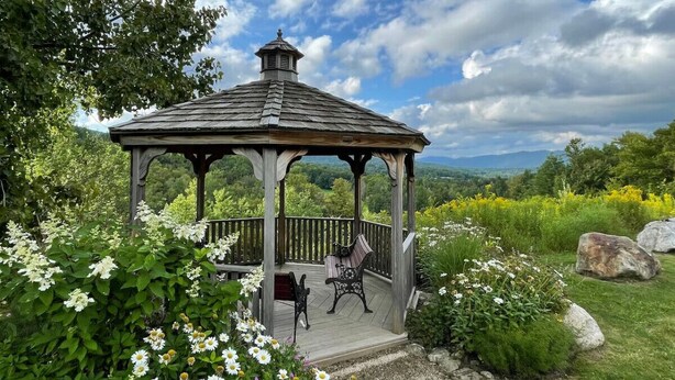A wooden gazebo with a roof, surrounded by lush greenery and flowers.