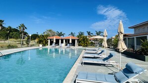 Une piscine entourée de palmiers, agrémentée de chaises longues et de parasols, et bordée d’un bâtiment au toit rouge.