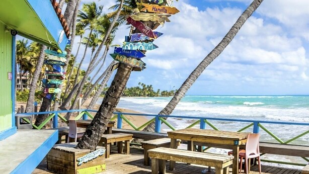 Une terrasse en bois avec des bancs, d’où l’on peut admirer une plage, des palmiers et un panneau indicateur indiquant différentes directions.