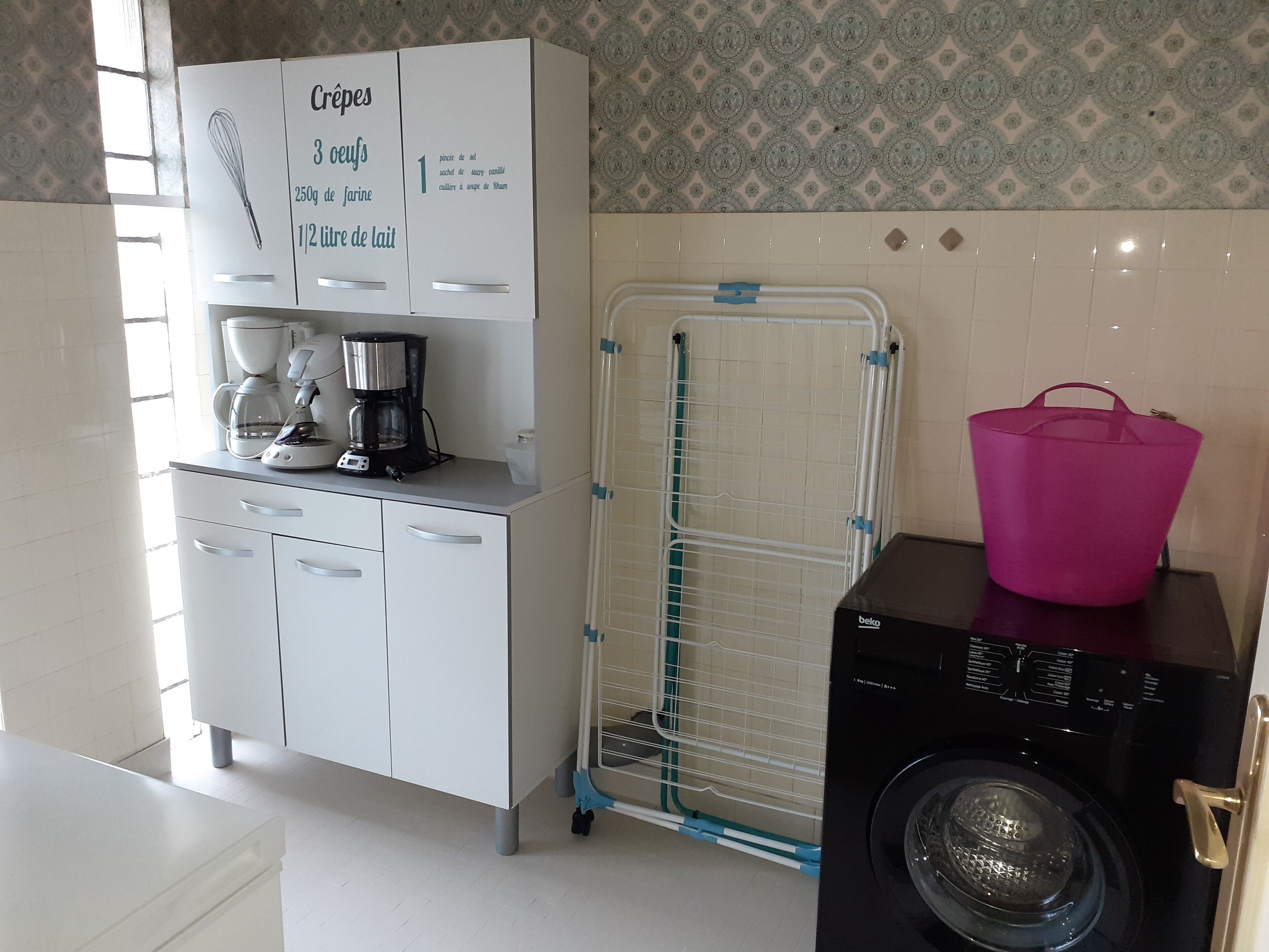Une cuisine équipée d’une machine à café, d’un lave-linge et d’un meuble blanc sur lequel est posée une carte de recettes.