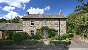 Une maison en pierre avec une porte grise, un petit jardin et une allée pavée.