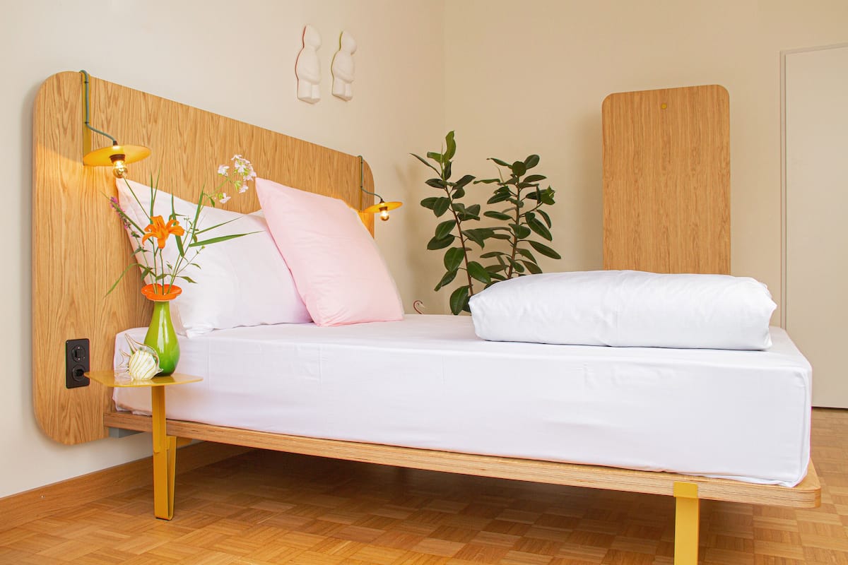 Een bed met een houten hoofdbord, een nachtkastje met een lamp en een potplant.