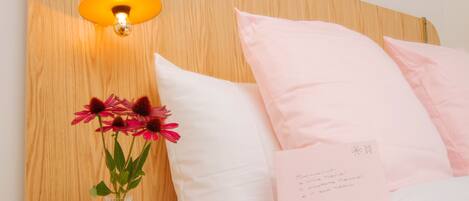 Una cama bien hecha con almohadillas rosas y una colcha blanca, un jarrón con flores rosadas y una lámpara fijada a la pared.