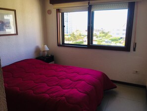 Une chambre à coucher comprenant un lit, une table de chevet, une lampe et une fenêtre donnant sur un immeuble et de la verdure.