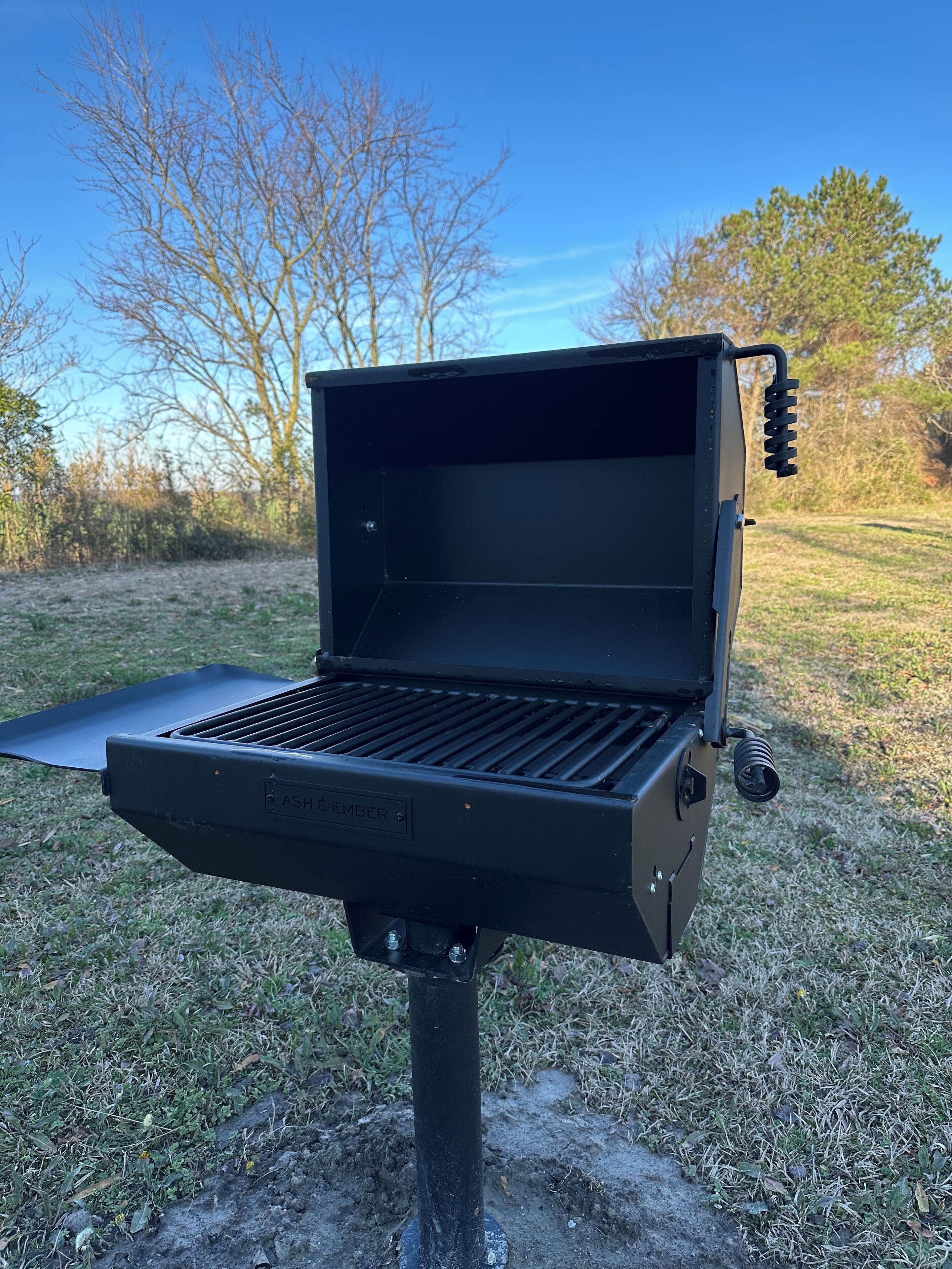 A portable grill with a flat cooking surface and a side shelf, set on a grassy area with trees in the background.