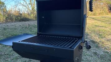 A portable grill with a flat cooking surface and a side shelf, set on a grassy area with trees in the background.