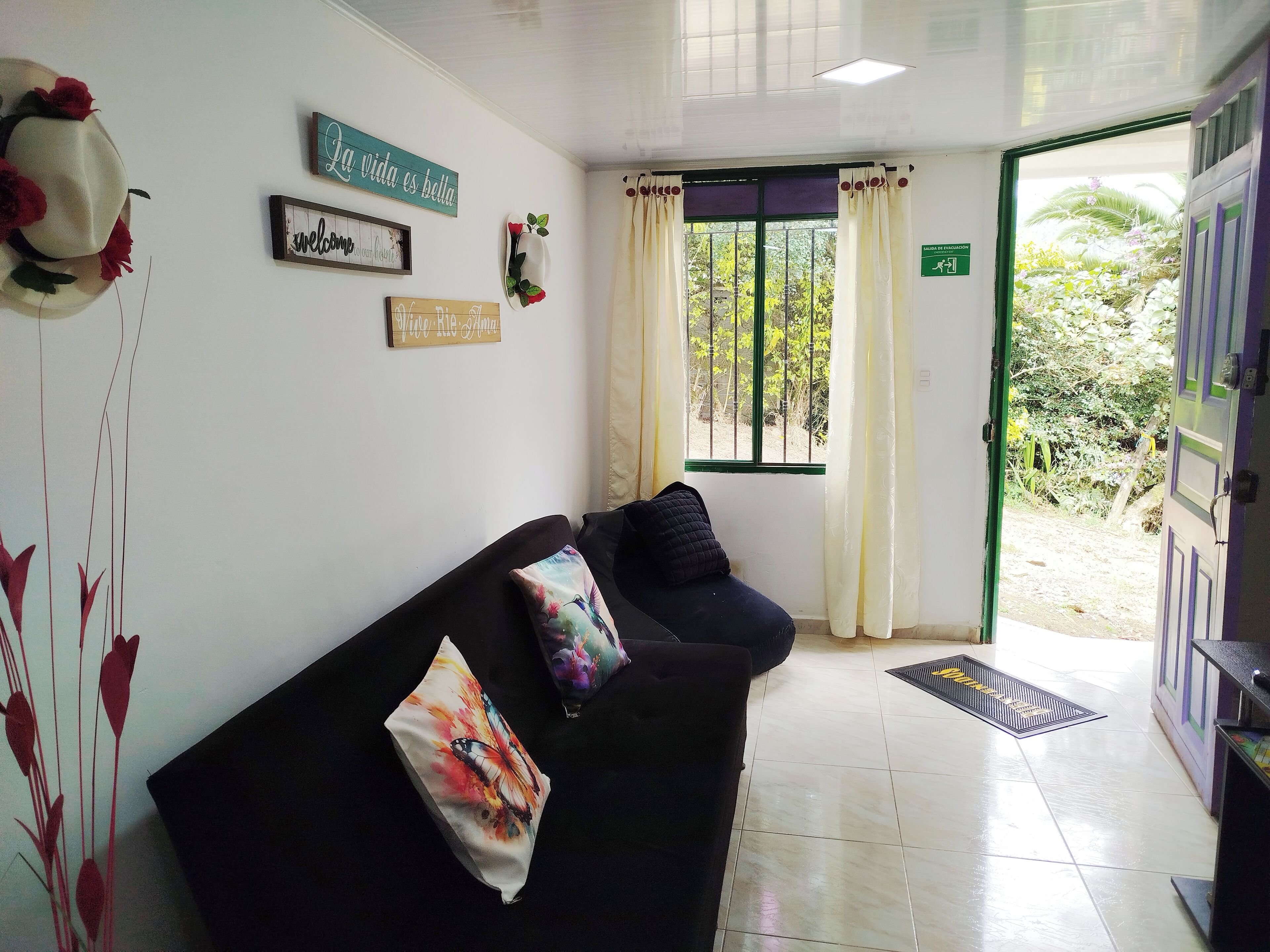 Una zona de recepción con un sofá negro, un muro decorativo, una ventana con cortinas y una puerta que da al exterior.