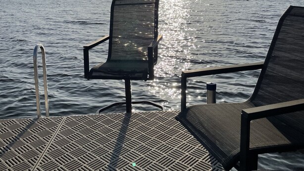 A deck with two lounge chairs facing a calm lake, a metal ladder, and a metal railing.