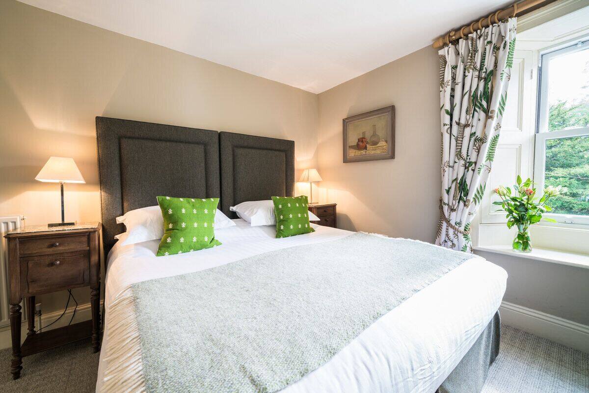 A neatly made bed with a grey and white duvet, two green pillows, and a headboard with a dark panel. There are two bedside tables with lamps, a window with curtains, and a framed picture on the wall.
