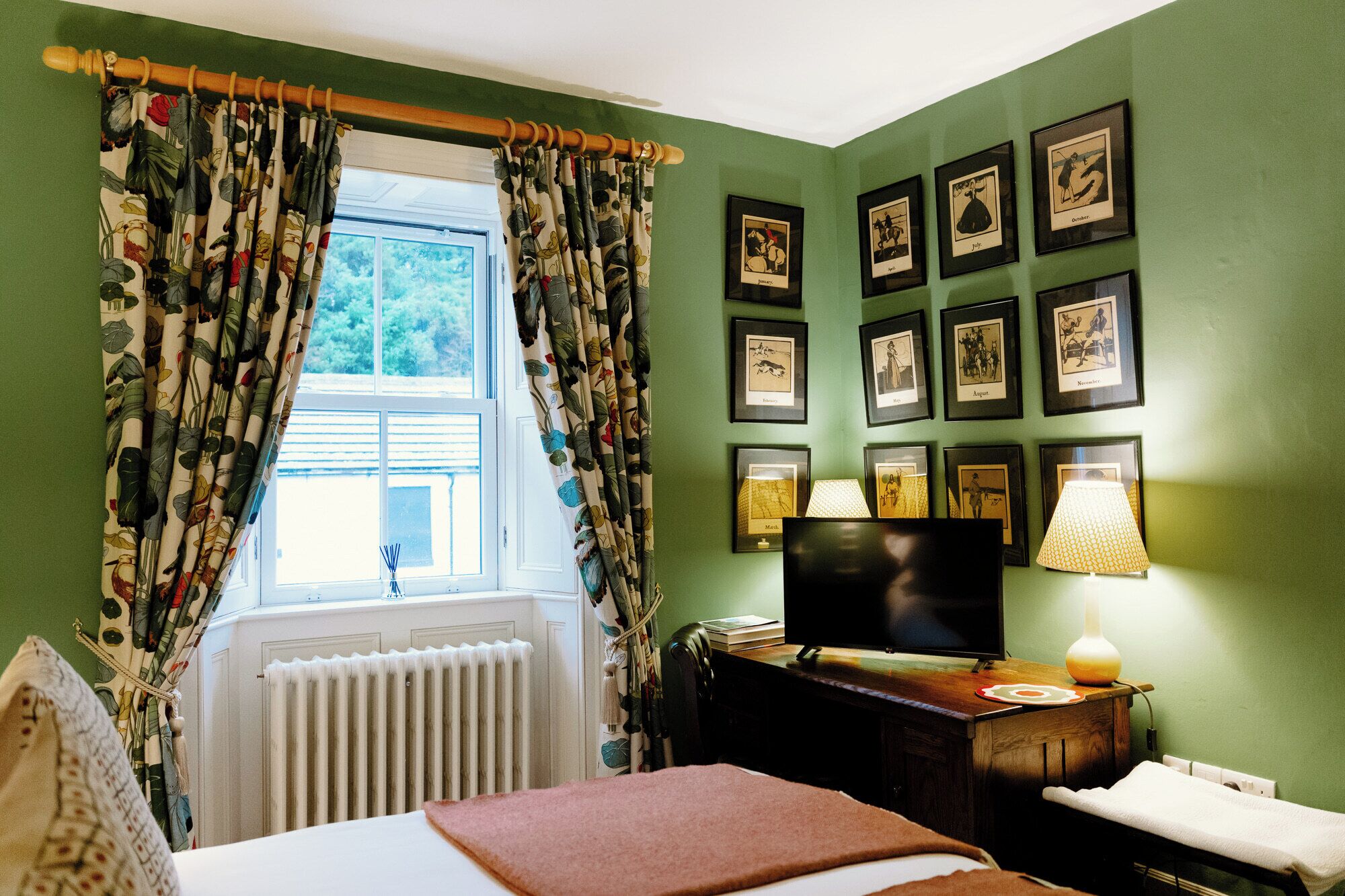 A room with a bed, a television, a lamp, and a wall with framed pictures.
