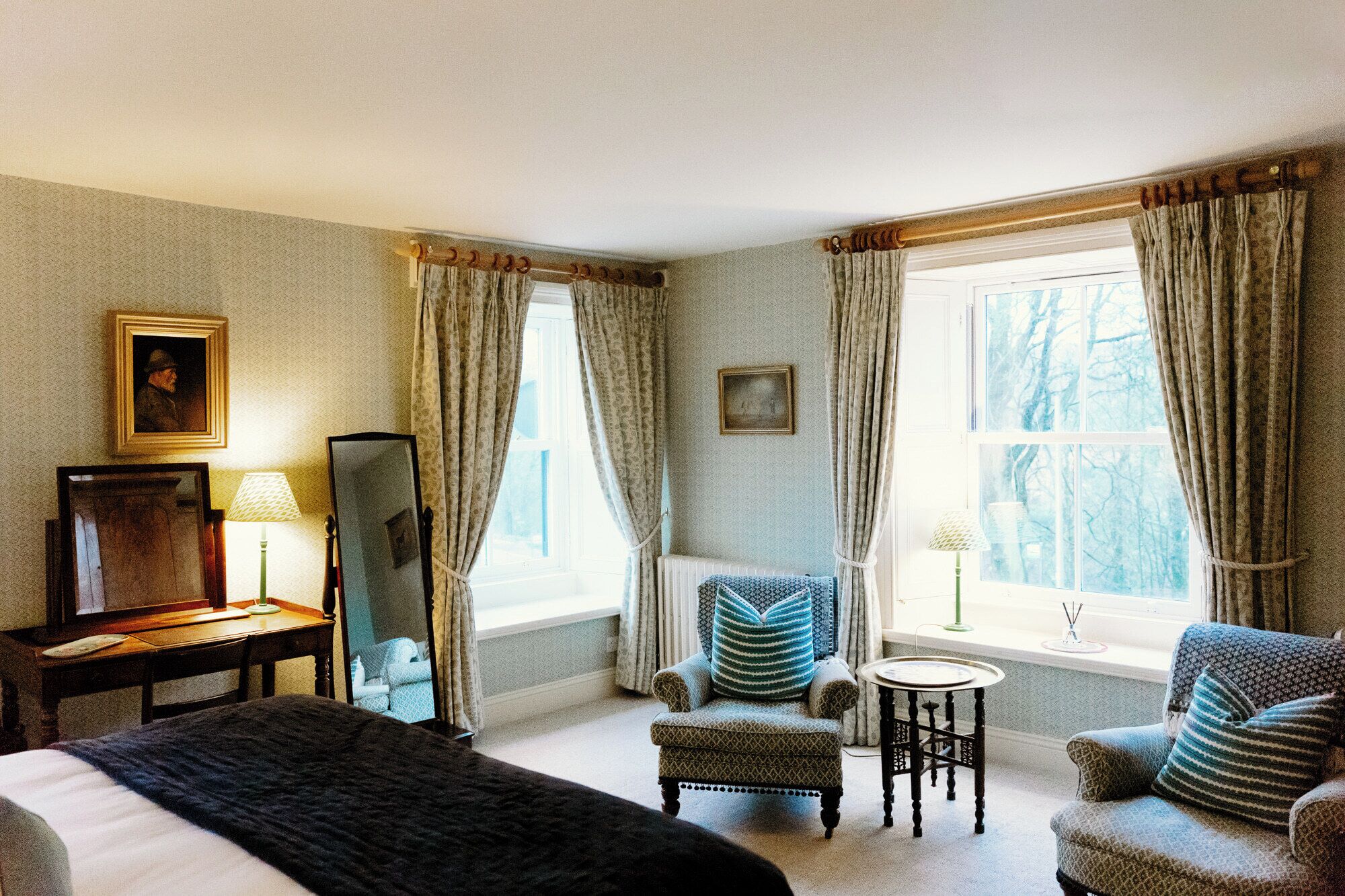 A hotel room with a bed, two armchairs, a small table, a mirror, and a window with curtains.
