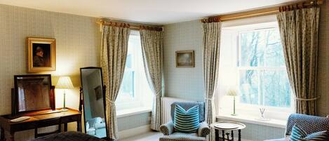 A hotel room with a bed, two armchairs, a small table, a mirror, and a window with curtains.