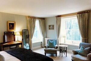 Quarto de hotel com cama, duas poltronas, uma mesinha, espelho e janela com cortinas.