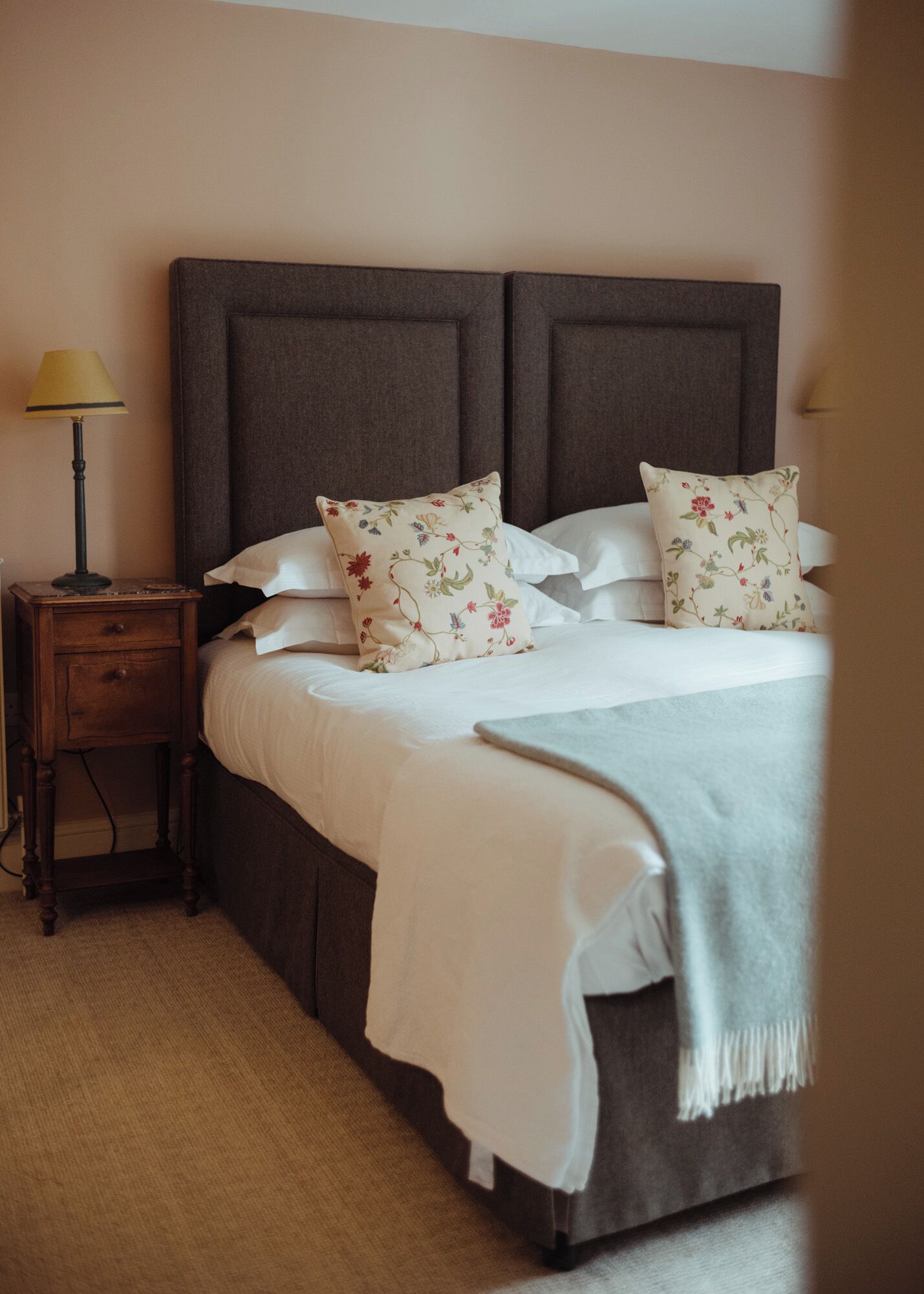 A neatly made bed with floral pillows and a white bedspread, a small wooden nightstand with a lamp, and a plain wall.