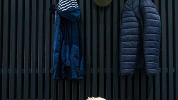Un perro sentado en un banco de madera, con un sombrero de rayas verticales y una chaqueta azul colgada en la pared.