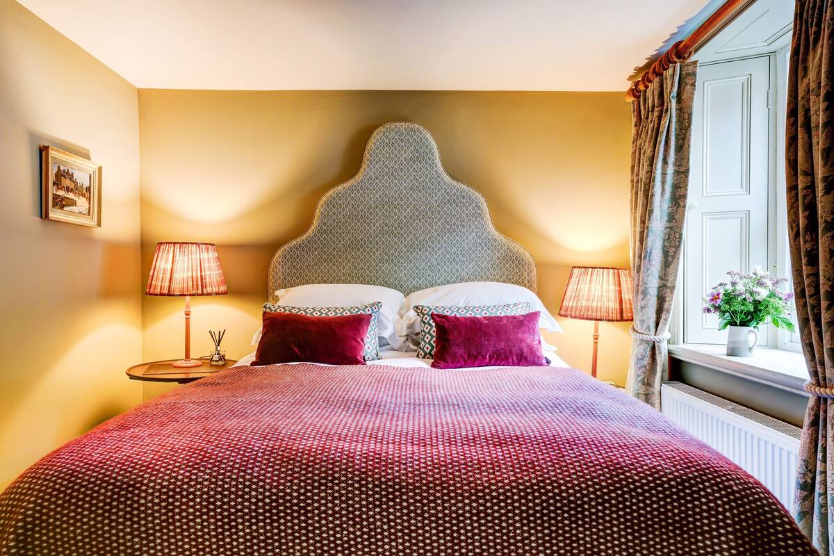 A hotel room with a large bed, two bedside lamps, a headboard, and a window with curtains.