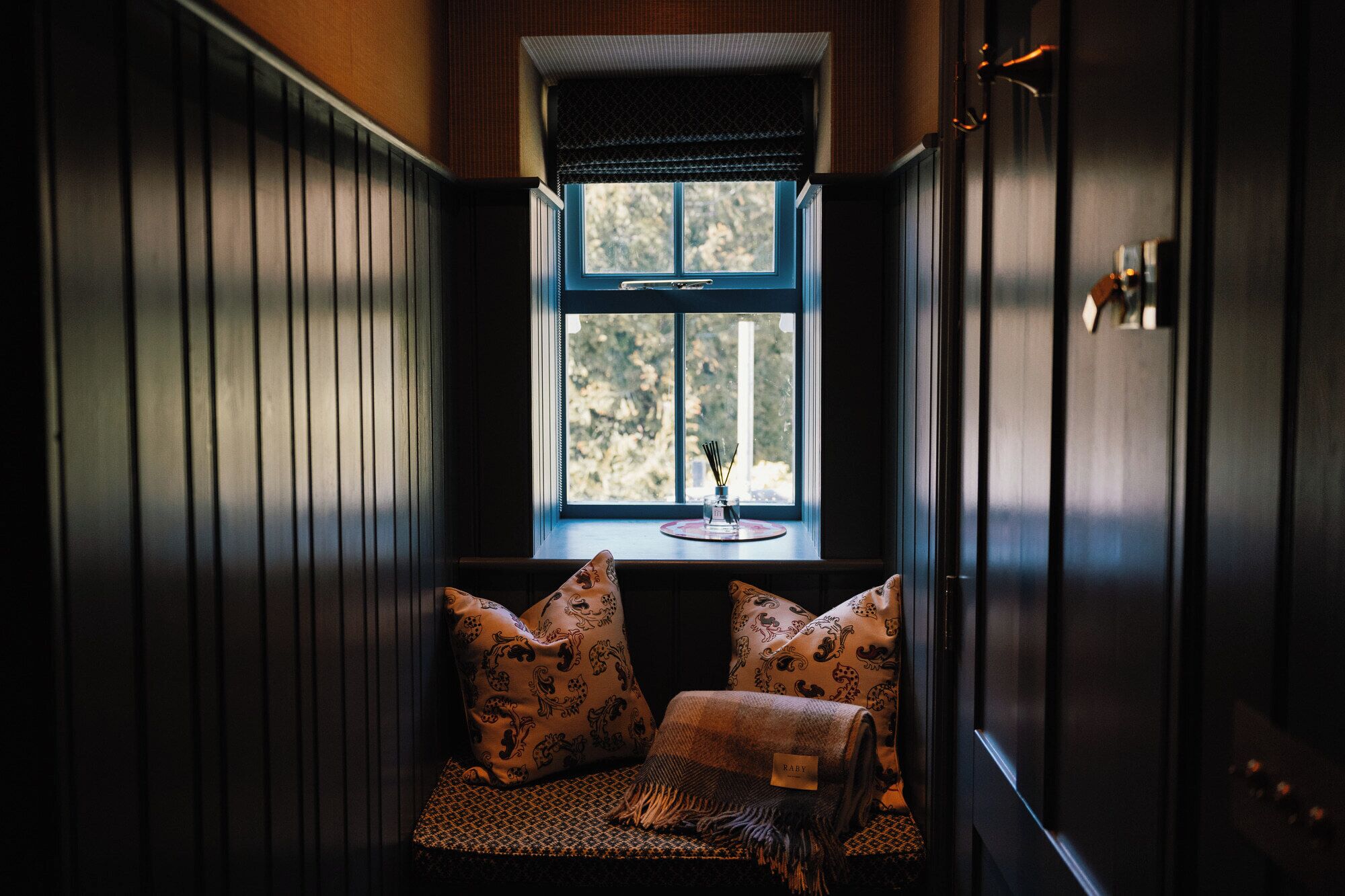 A cozy seating area with patterned cushions and a blanket, a window with a view of trees, and a small table with a candle.