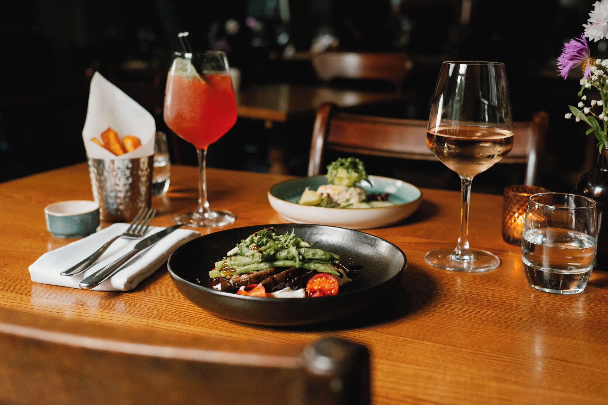 A dining table with a plate of food, a glass of white wine, a cocktail, and a small bowl of fries.