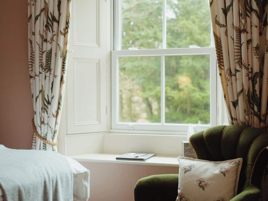 Ein Zimmer mit Fenster, ein grüner Sessel mit Blumenkissen und ein weißes Bett mit blauer Decke.