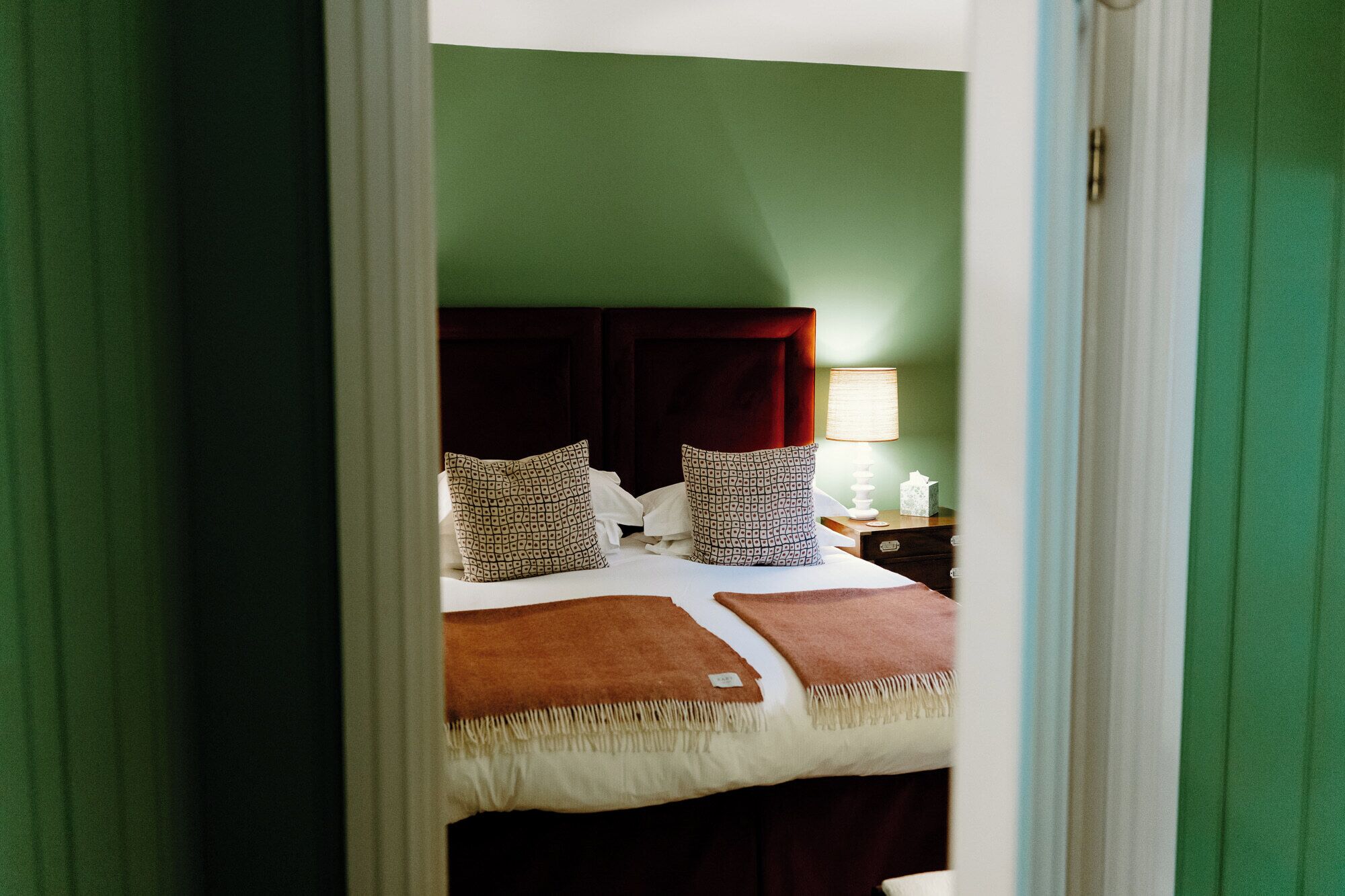 Quarto de hotel com cama, almofadas, mesa de cabeceira com abajur e criado-mudo.