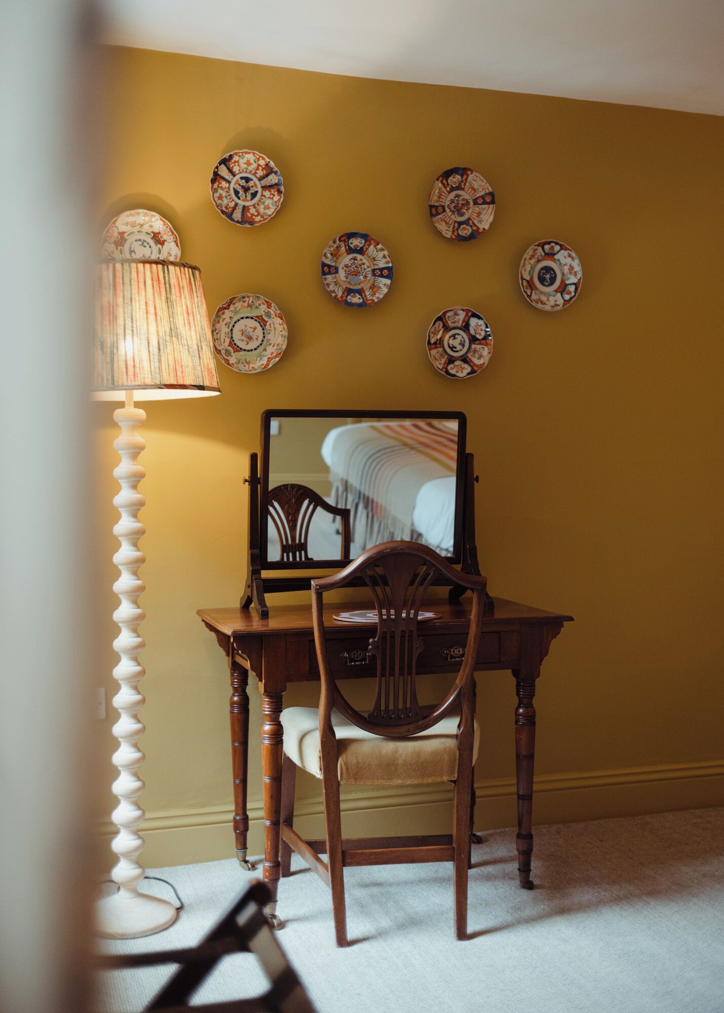 Una habitación con un escritorio de madera, una silla, una lámpara y un espejo en la pared.