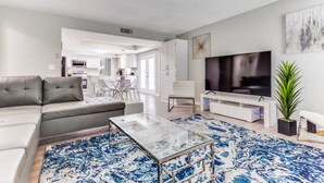 A modern living room with a sofa, coffee table, and TV. There is a kitchen in the background. - Reston NEW Bright, Airy Home w/ In Ground Pool & Grill (Richardson)