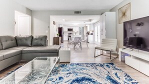 A modern living room with a sectional sofa, a glass coffee table, and a TV on a white stand. - Reston NEW Bright, Airy Home w/ In Ground Pool & Grill (Richardson)