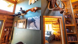 A rustic cabin interior with wooden walls, a ceiling with wooden beams, mounted animal heads, framed photographs, and a bookshelf.