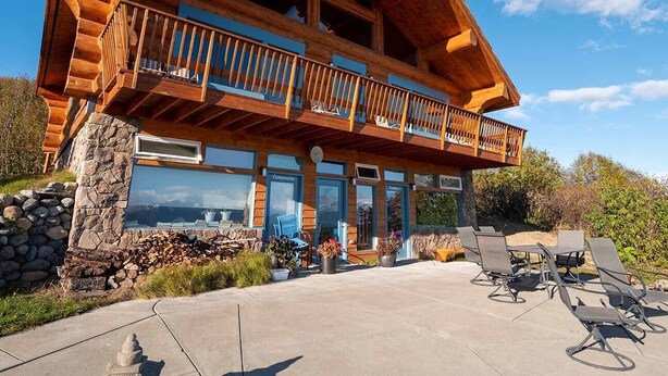 A log cabin with a balcony, stone foundation, outdoor seating, and a view of the landscape.
