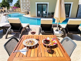 Zona de comedor junto a la piscina, con una mesa para dos, dos platos de comida y dos copas de vino tinto.