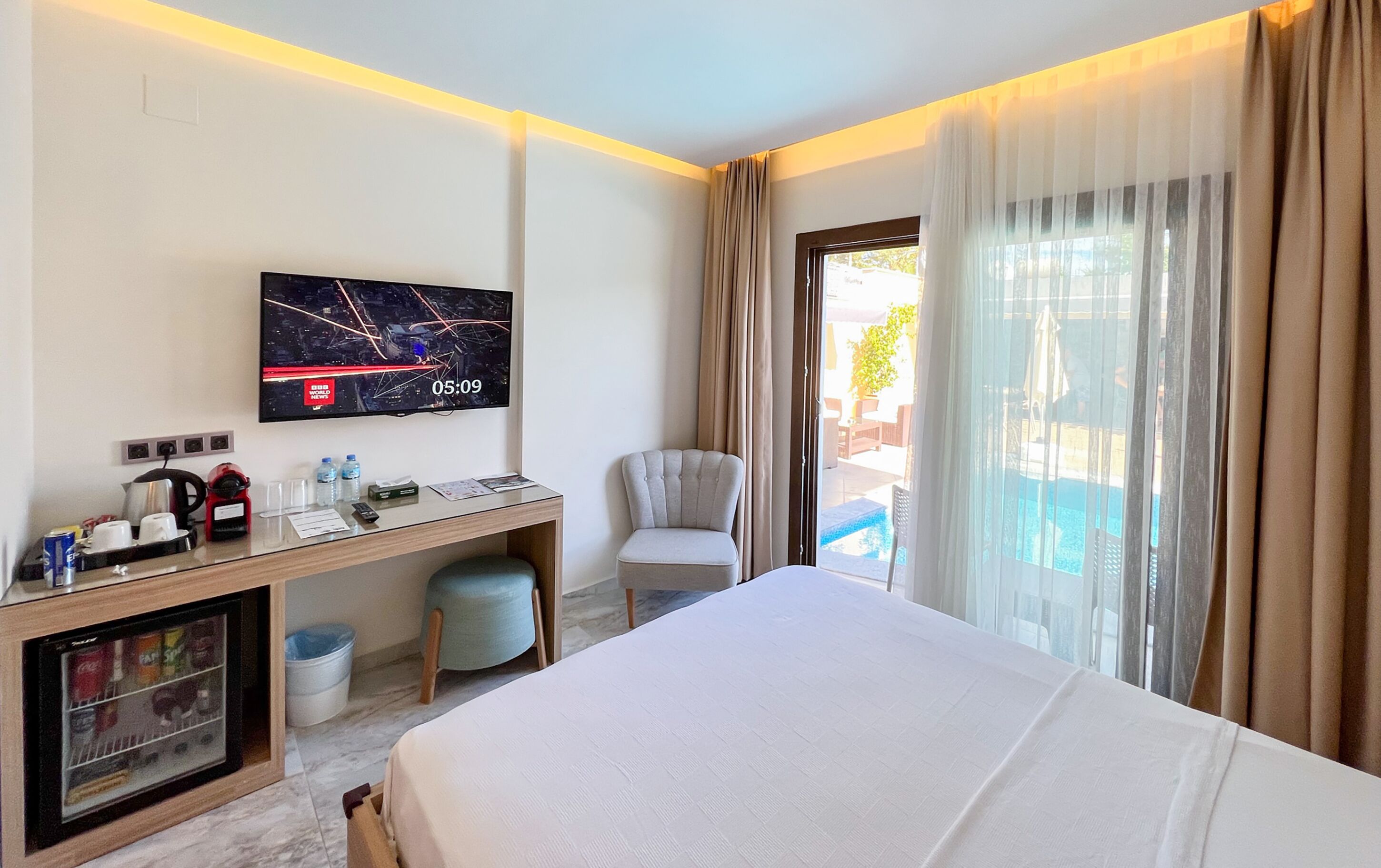 Habitación de hotel con cama, escritorio con televisor, silla, mesita con tetera, tazas y una botella de agua. Se ve una ventana con cortinas y una vista a la piscina.