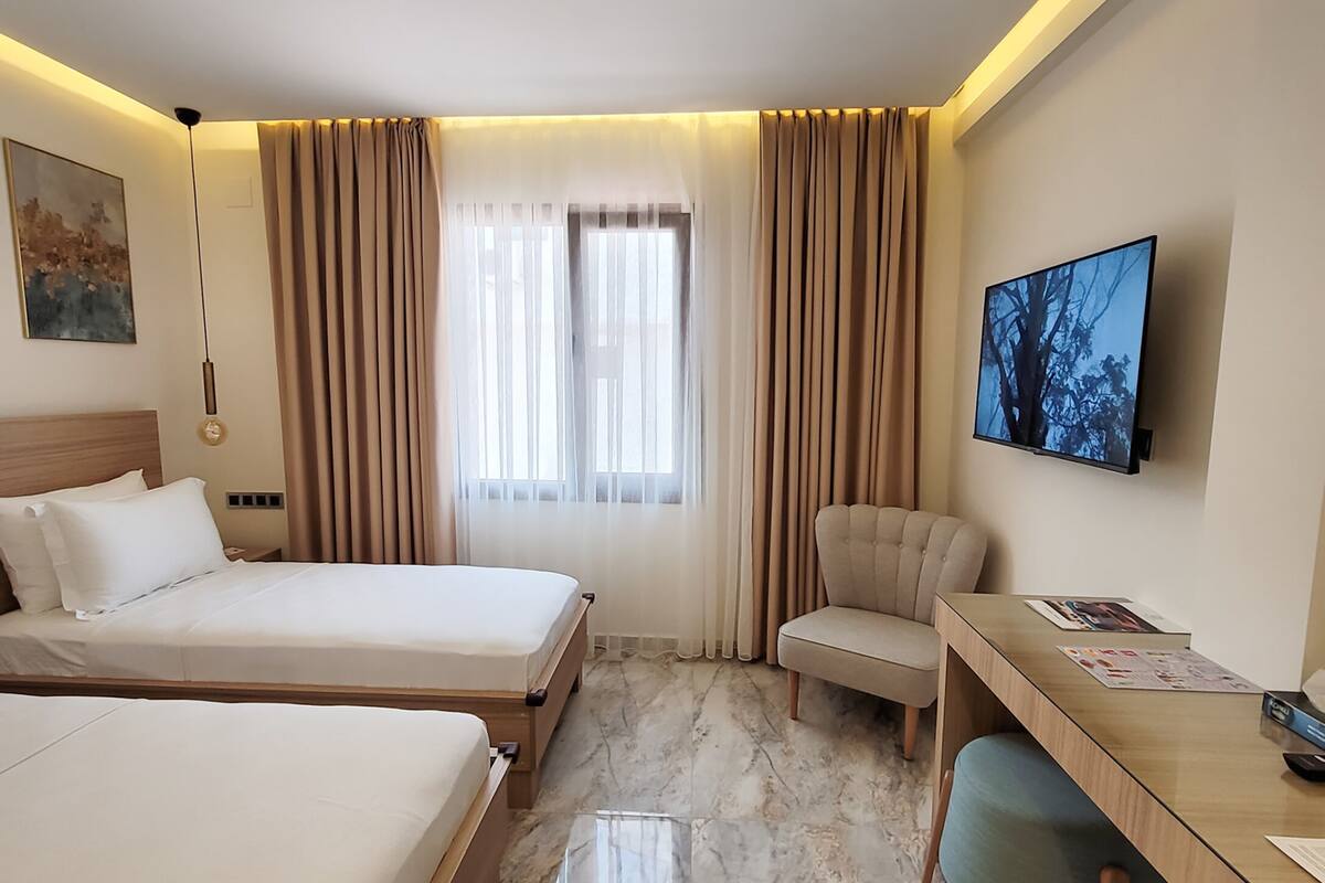 A hotel room with two beds, a desk, a chair, a TV, and a window with curtains.
