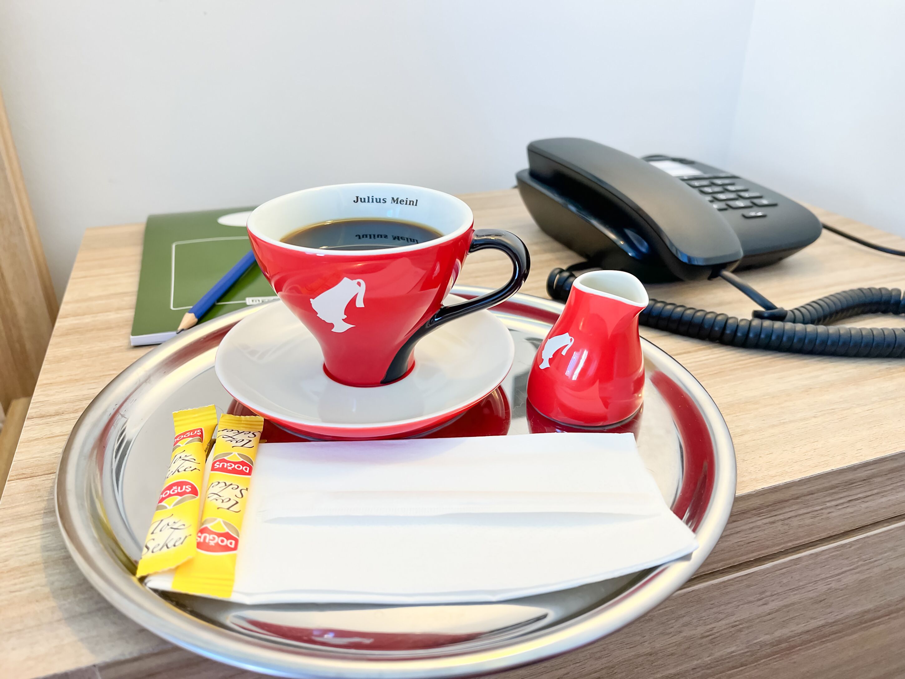 Ein Tablett mit einer roten Tasse mit dem Aufdruck "Julius Meinl", einer kleinen roten Kanne, einem Telefon, einem grünen Notizbuch, einem blauen Stift und zwei Teebeuteln "Drogus".
