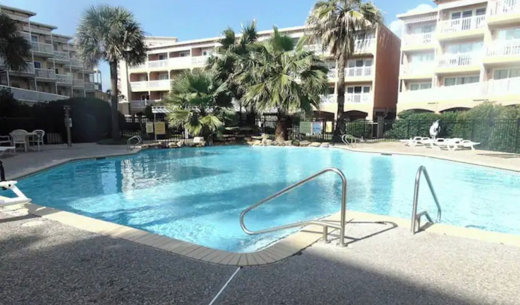 A swimming pool with a metal safety railing, surrounded by palm trees and a multi-story building with balconies.