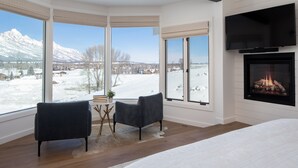 A bedroom with a large bed, a fireplace, a TV mounted above it, and a view of a snowy landscape through large windows. - Sweeping Teton views, modern renovation. Minutes from town & National Parks. (Jackson)