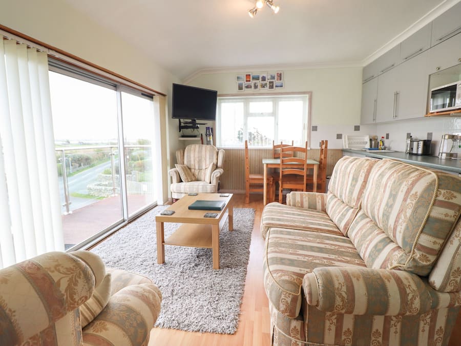 A living room with a sofa, armchairs, a coffee table, and a TV. There's a dining area with a table and chairs. The room has large windows and a view of the outside.