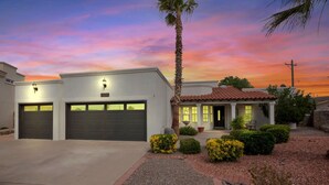 A two-story house with a garage, a small porch with a table and chairs, and a well-maintained garden with shrubs and a palm tree. - Incredible home with custom pool and fountain (El Paso)